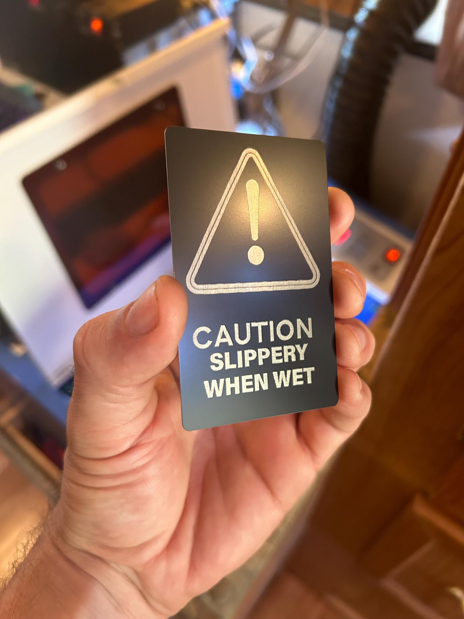 Hand holding a metal black/silver warning sign with 'Caution Slippery When Wet' text.