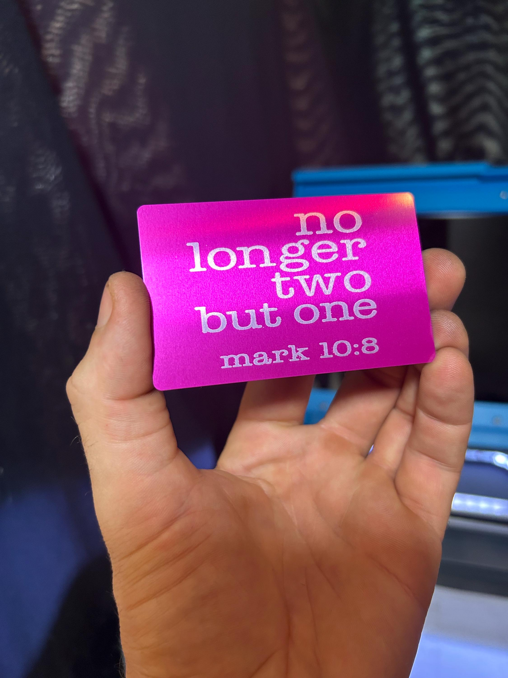 Hand holding a metal purple and silver laser engraved keepsake card with text 'no longer two but one' from Mark 10:8