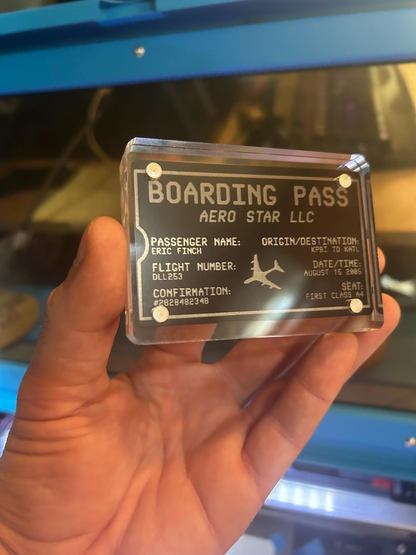 Hand holding a transparent acrylic display case with a metal laser engraved keepsake inside reading 'Boarding Pass' text and branding