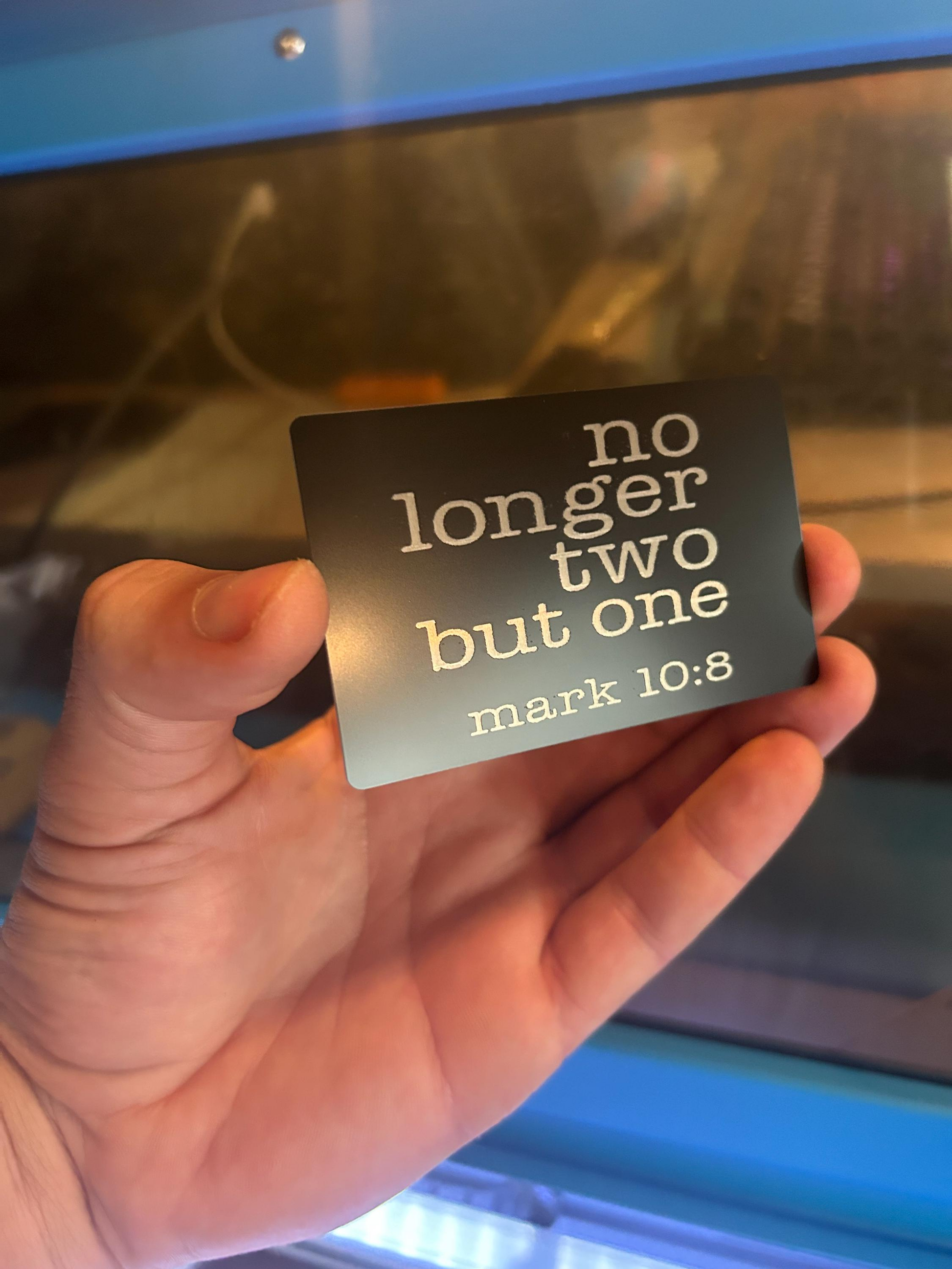 Hand holding a metal inspirational keepsake card with 'no longer two but one' and 'mark 10:8' text.