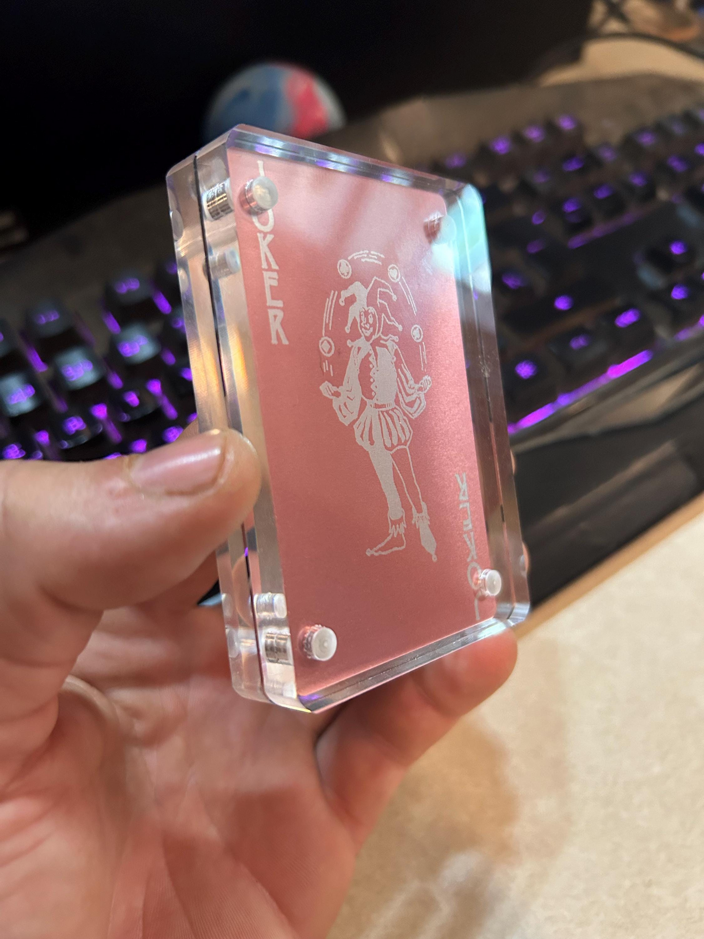 Clear card holder with engraved design held by a hand, blurred keyboard in the background