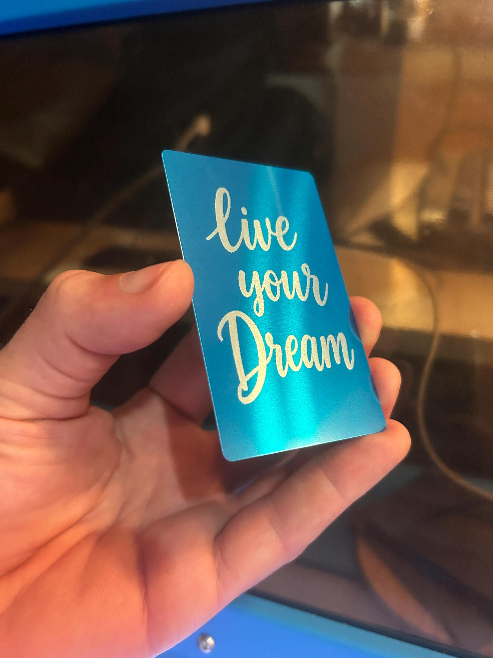 Hand holding a blue and silver metal keepsake card engraved with “Live Your Dream” typography text design
