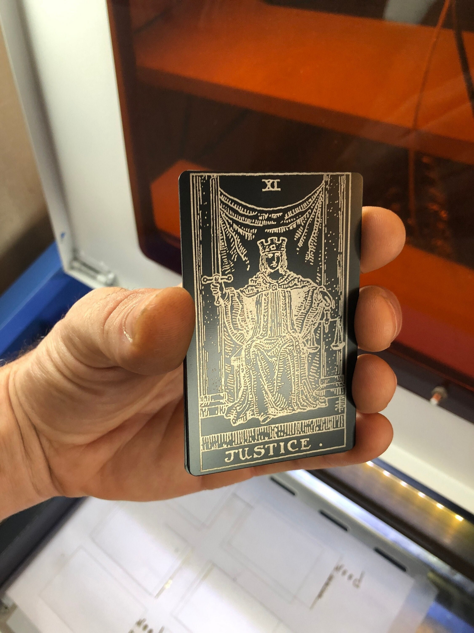 Hand holding a laser engraved anodized aluminum tarot card with 'Justice' inscribed, blurred background