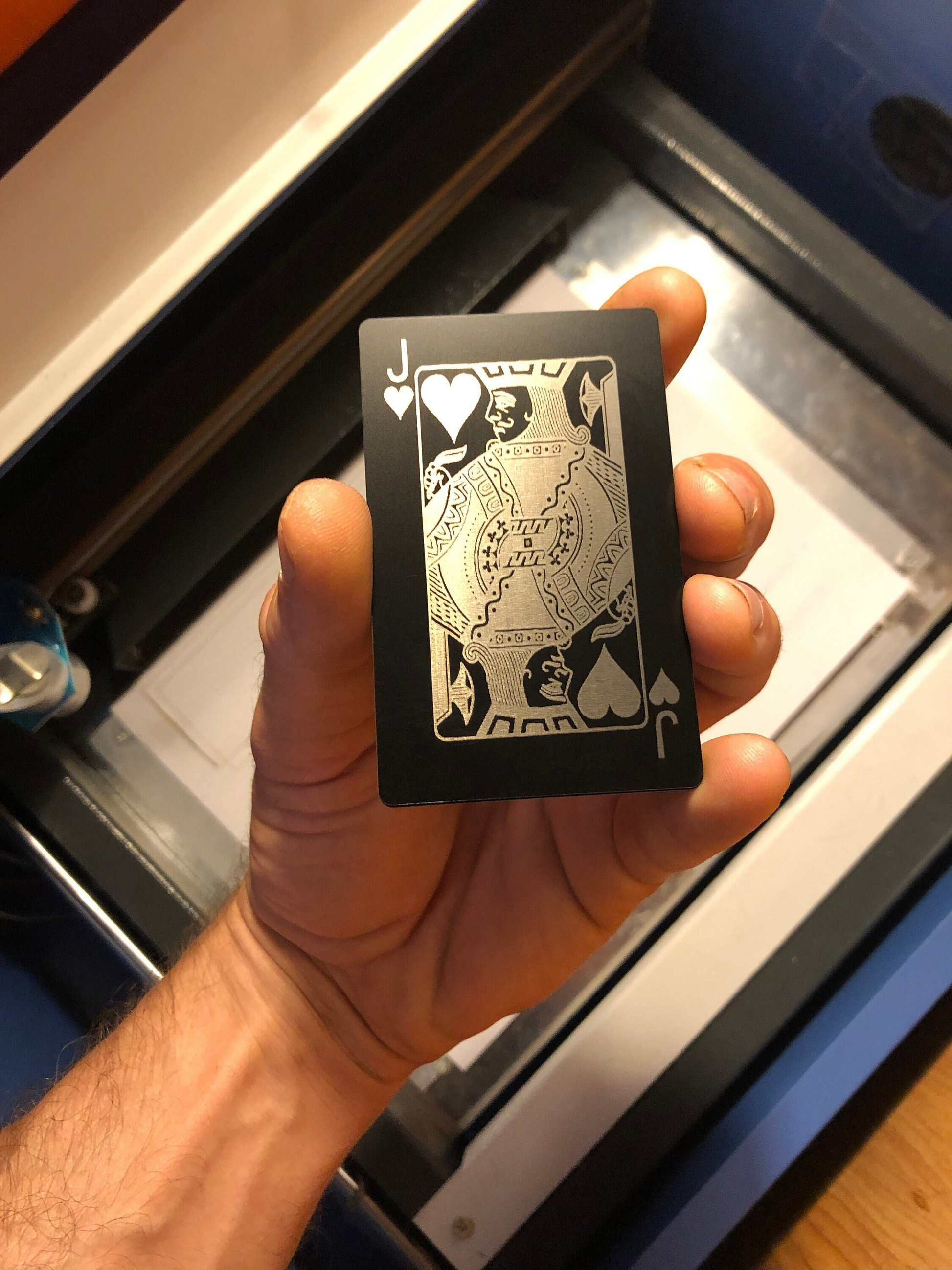 Hand holding a metal black/silver Jack of Hearts poker card in front of a machine background