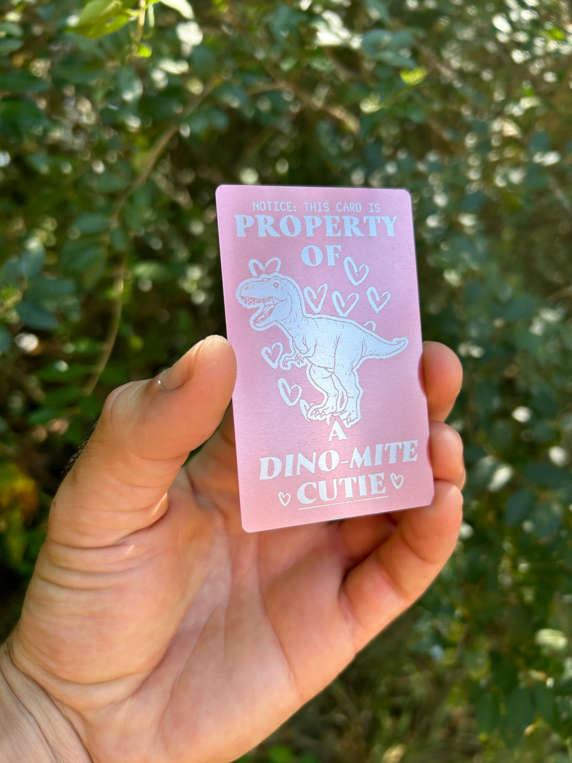 Hand holding a cute metal pink card with a T Rex design and text against a green leafy background