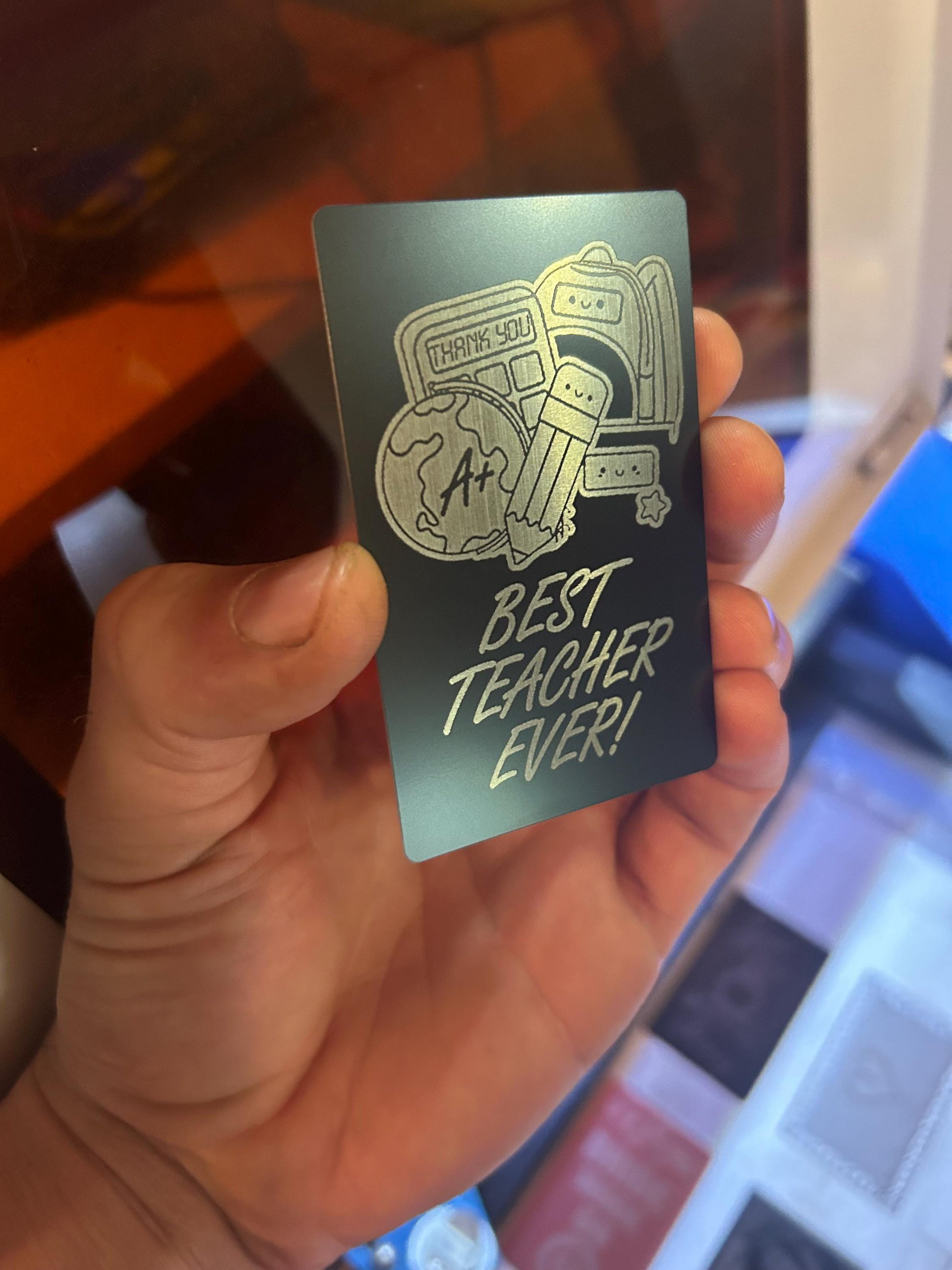 Hand holding a metal gift keepsake card with 'Best Teacher Ever!' text and graphics.