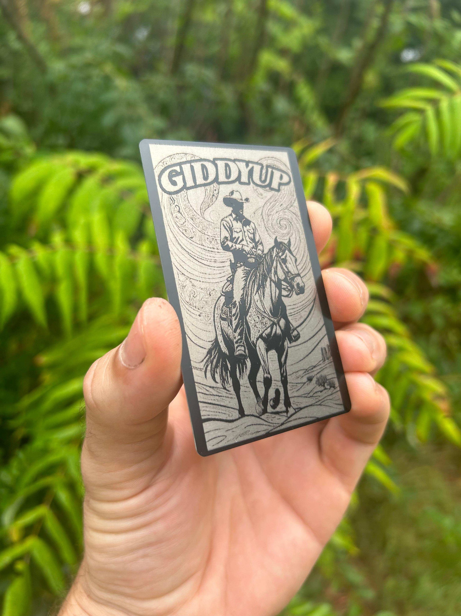Hand holding a metal Cowboy 'Giddy Up' card with a horse and rider illustration against a green background