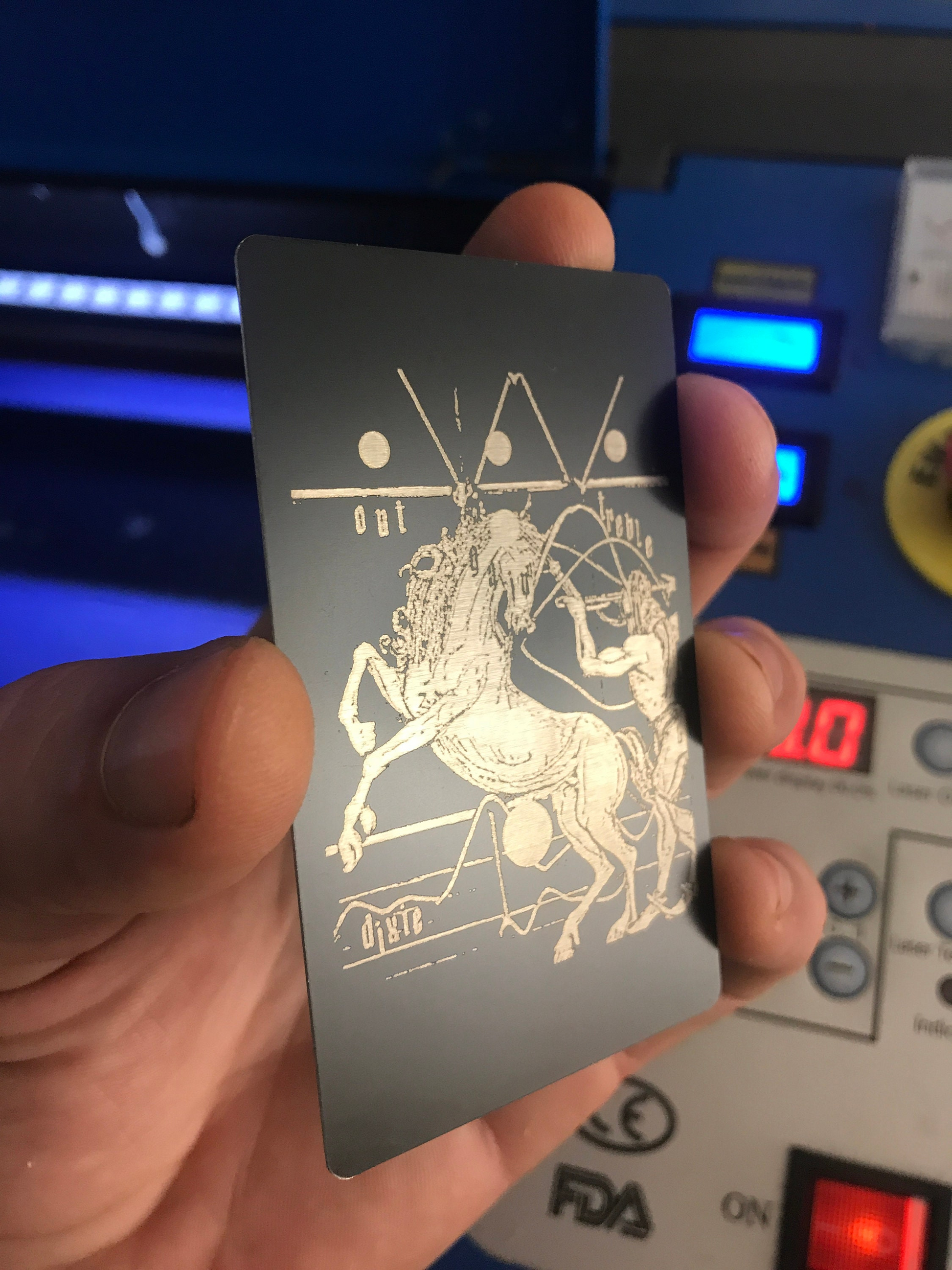 Hand holding a black and silver metal card card with an astrological design against a blurred machine background