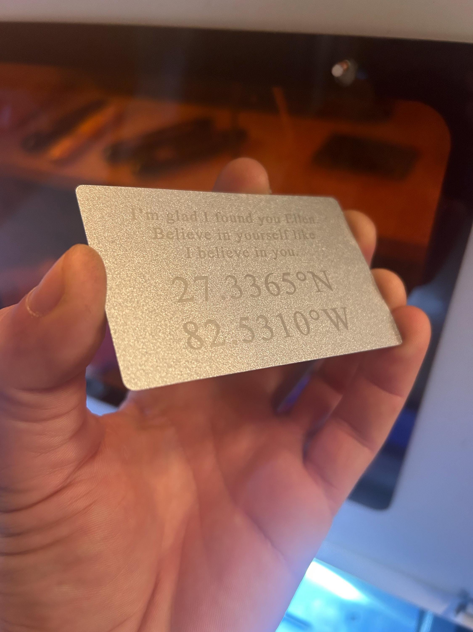 Hand holding a silver-colored card with personalized GPS coordinates and text "I'm Glad I Found You Chris, Believe In Yourself Like I Believe In You"