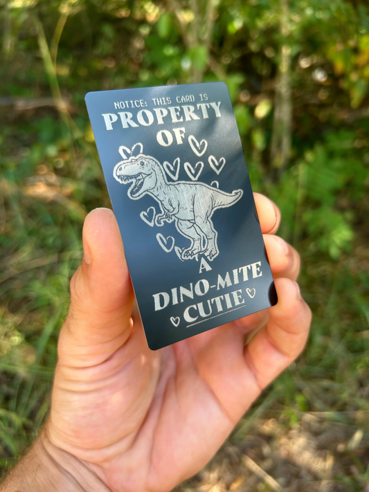 Hand holding a metal keepsake gift card with a cute sarcastic dinosaur illustration and text in an outdoor setting