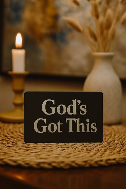 Decorative metal placard with 'God's Got This' text on a table with a candle and vase in the background.