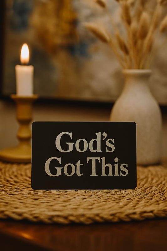 Decorative metal placard with 'God's Got This' text on a table with a candle and vase in the background.