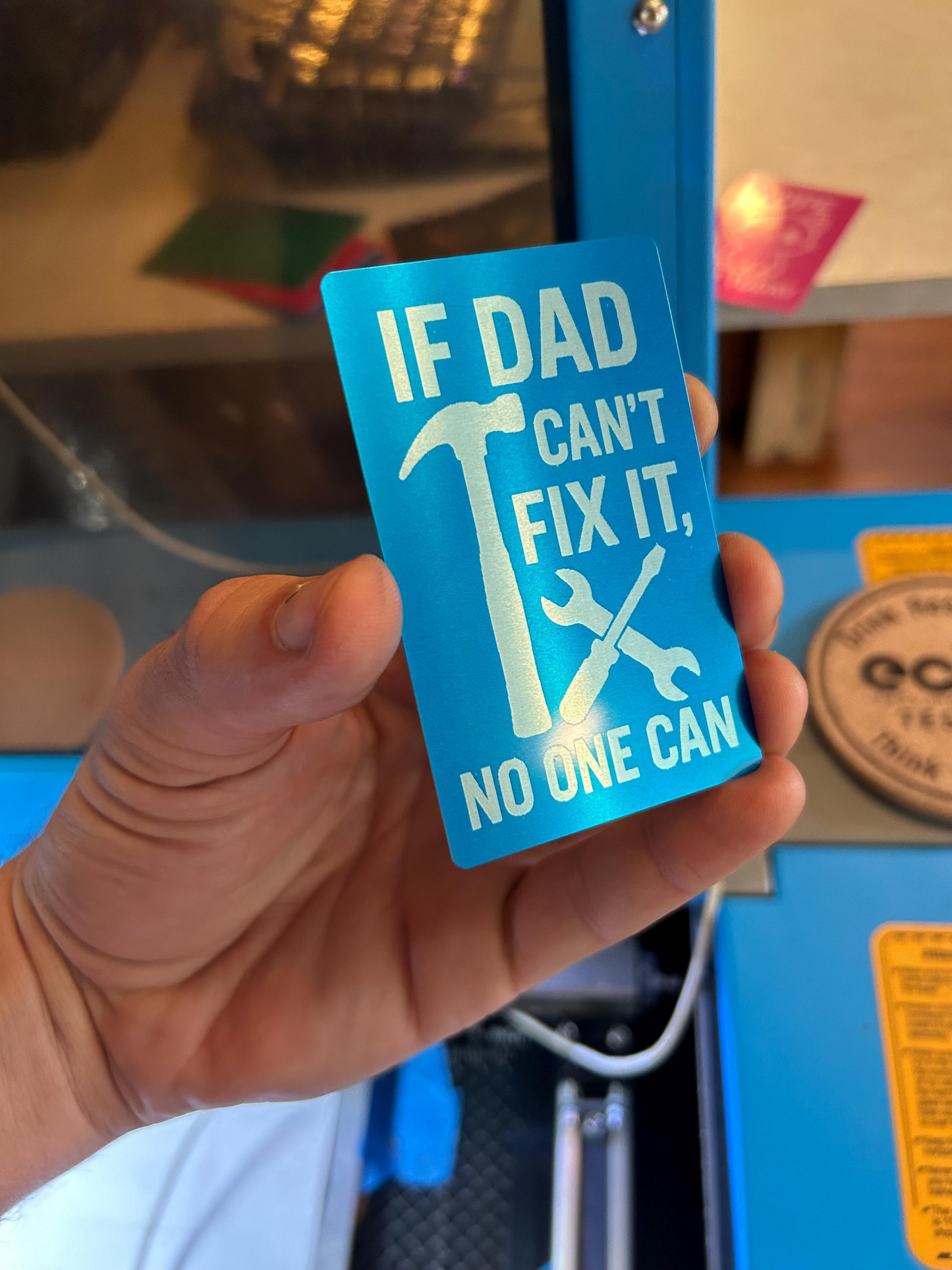 Hand holding a laser engraved metal blue/silver keepsake wallet insert with text and tools graphics, set against a blurred indoor background.