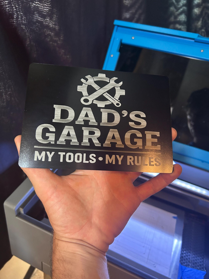 Hand holding a metal shop sign with 'Dad's Garage, My Tools, My Rules' text.
