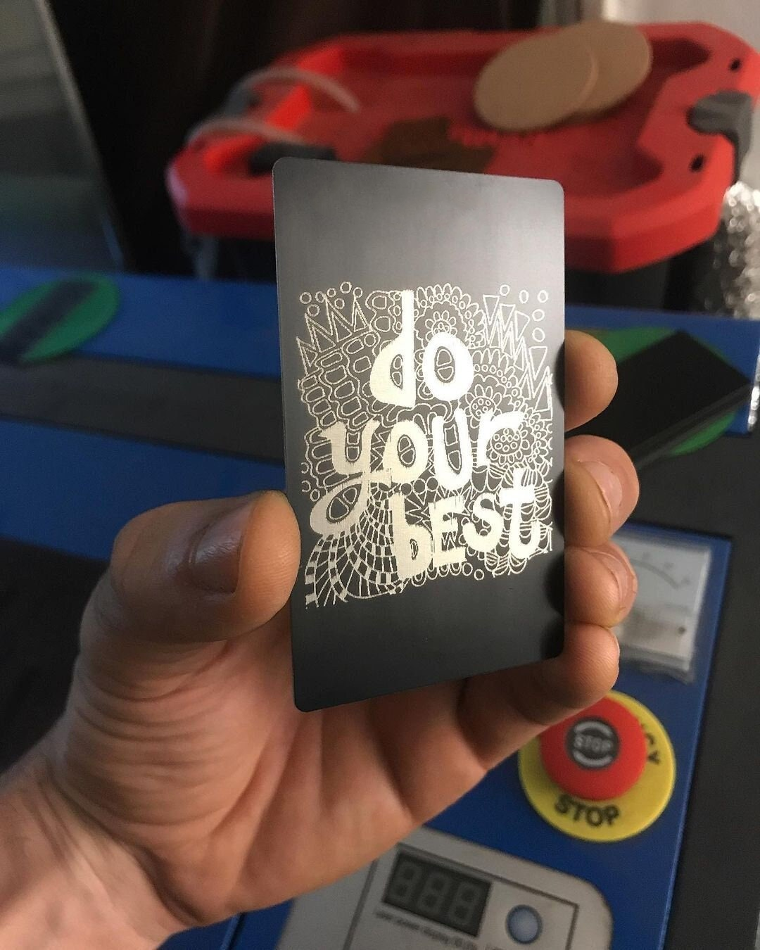 Hand holding a black metal gift card with 'Do Your Best' text in front of a workshop background