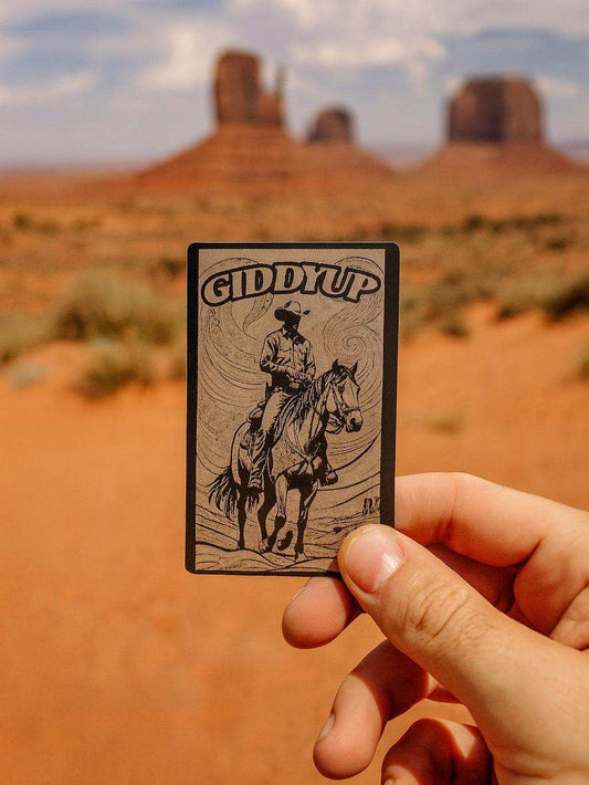 Hand holding a card with 'Giddy Up' text and a cowboy illustration against a desert landscape.