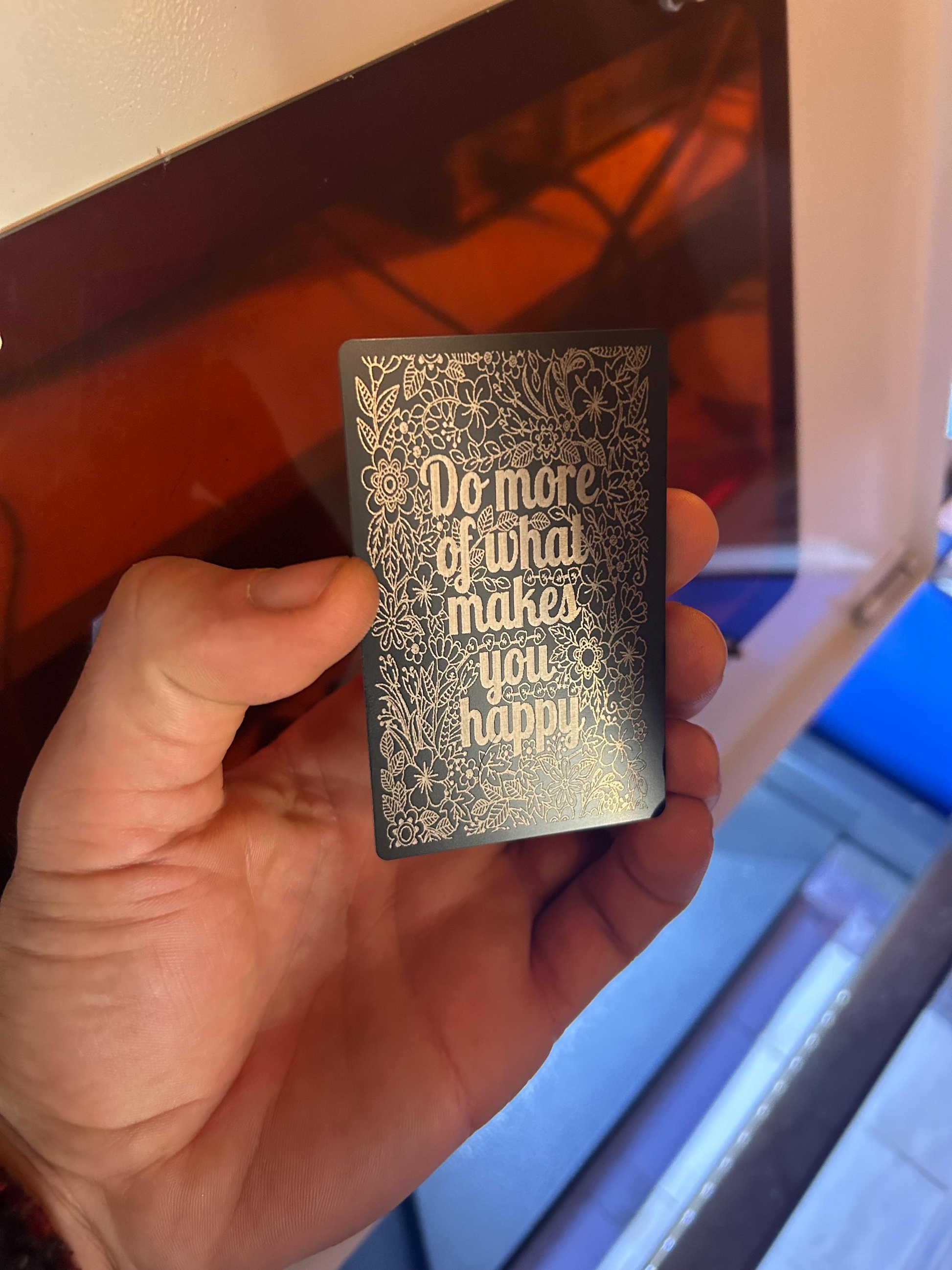 Hand holding a small black anodized aluminum laser engraved keepsake with silver text that reads "Do More Of What Makes You Happy" with a floral graphic pattern against a laser engraving machine background