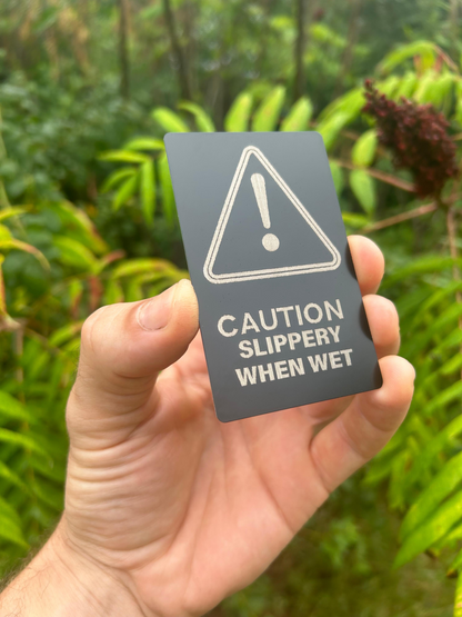 Hand holding a laser engraved metal signage placard with 'Caution Slippery When Wet' text in anodized aluminum with a green, natural background