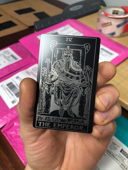 Hand holding a tarot card titled 'The Emperor' against several mailing packages on a blurred background.