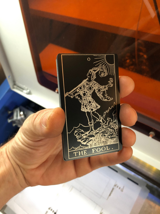Hand holding a metal black/silver tarot card engraved with 'The Fool' in front of a 3D printer.