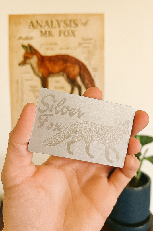 Hand holding an anodized aluminum card with 'Silver Fox' engraving in front of a fox-themed poster.
