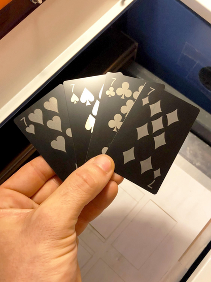 Hand holding metal playing cards with seven of hearts, seven of spades, 7 of clubs, 7 of diamonds laser engraved silver symbols in matte black against a blurred background