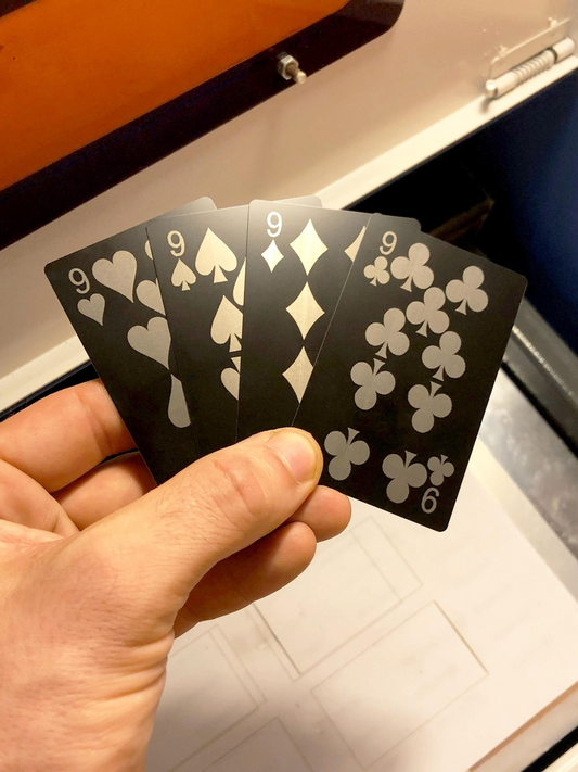 Hand holding 4 metal 9 playing cards with nine of hearts, nine of spades, nine of diamonds, nine of clubs, laser engraved in anodized aluminum in front of a laser machine