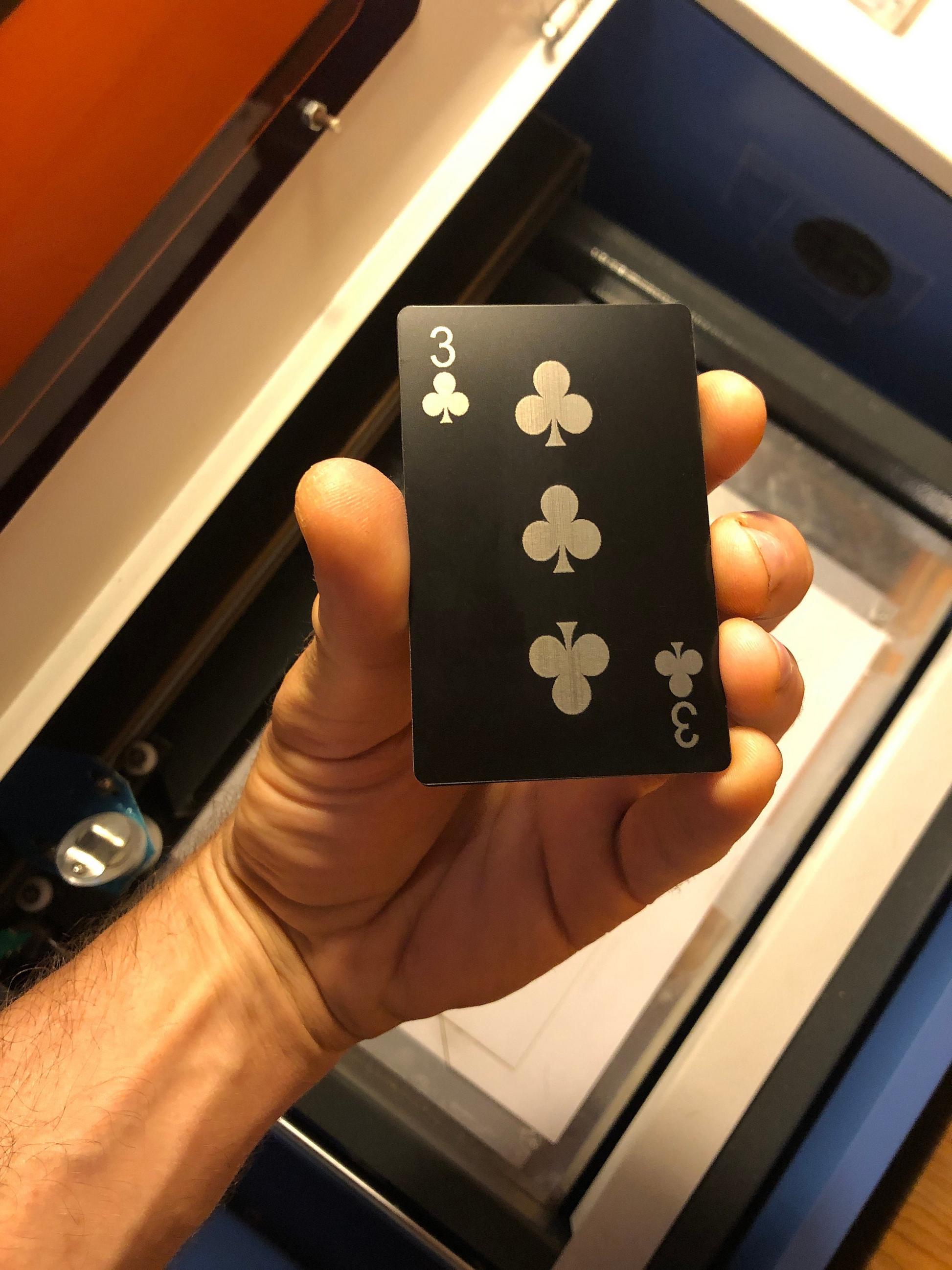 Hand holding a matte black playing card with three of clubs laser engraved on anodized aluminum, against a blurred background