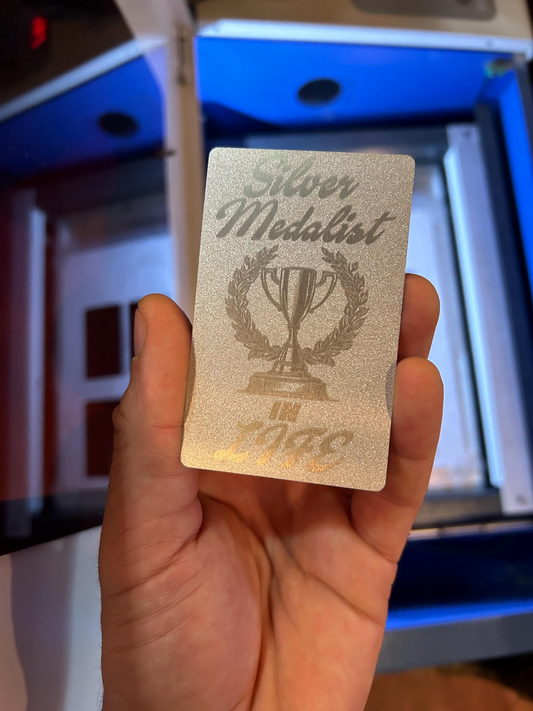 Hand holding a laser engraved novelty gag gift card that says "Silver Medalist In Life" with ironic trophy art, demotivational gift card