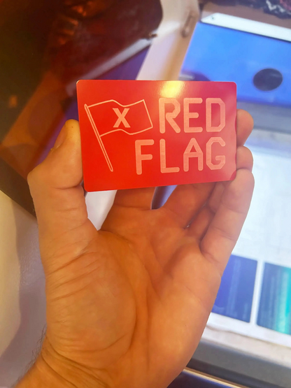 Hand holding a laser engraved novelty red card with 'X RED FLAG' text and flag symbol.