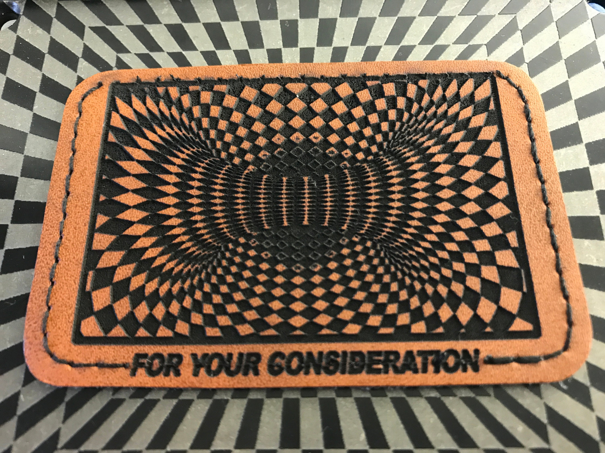 Brown leather patch with a geometric pattern and 'For Your Consideration' text on a checkered background