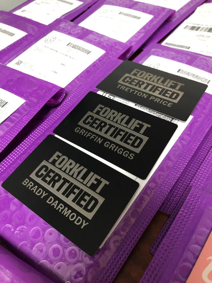 Metal Forklift Certified badges with personalized name engravings in black/silver anodized aluminum pictured on top of purple mailers.