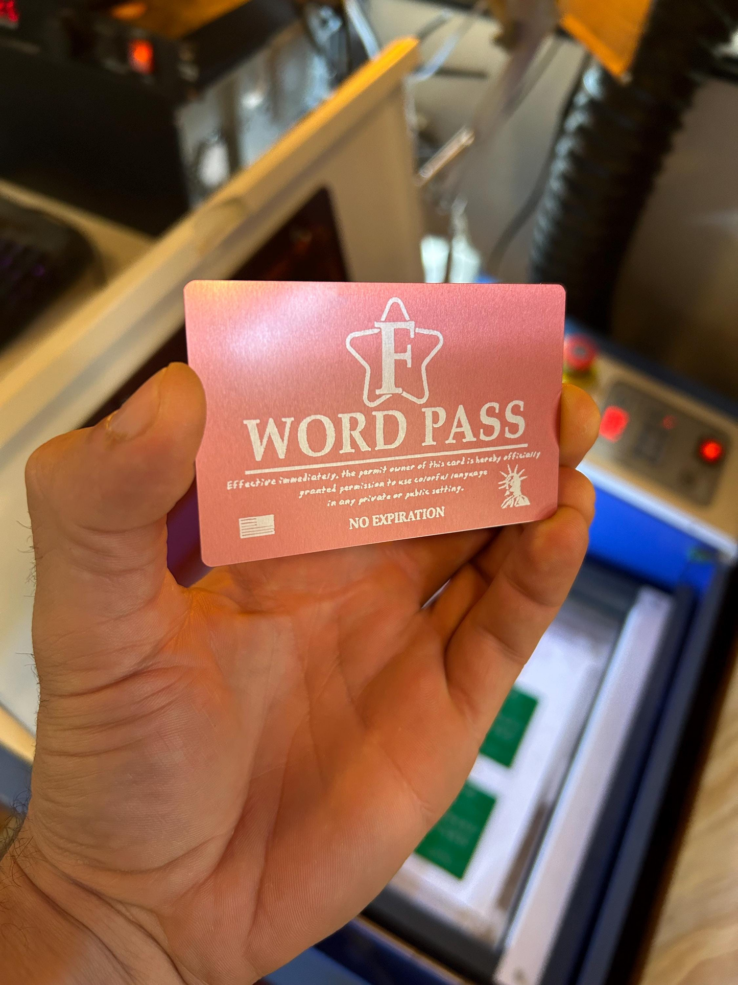 Hand holding a rose gold metal 'F WORD PASS' gag gift card in front of a machine.
