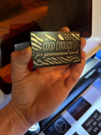 Hand holding a novelty metal gift card with 'Good Enough for Government Work' text and american flag pattern.