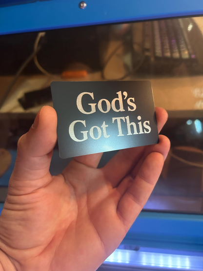 Hand holding a metal novelty keepsake card with 'God's Got This' laser engraved text in front of a computer screen.
