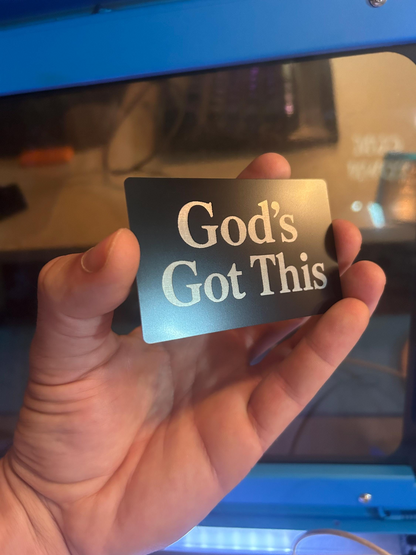 Hand holding a laser engraved metal business card with 'God's Got This' text in front of a blurred background