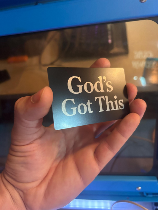 Hand holding a laser engraved metal business card with 'God's Got This' text in front of a blurred background