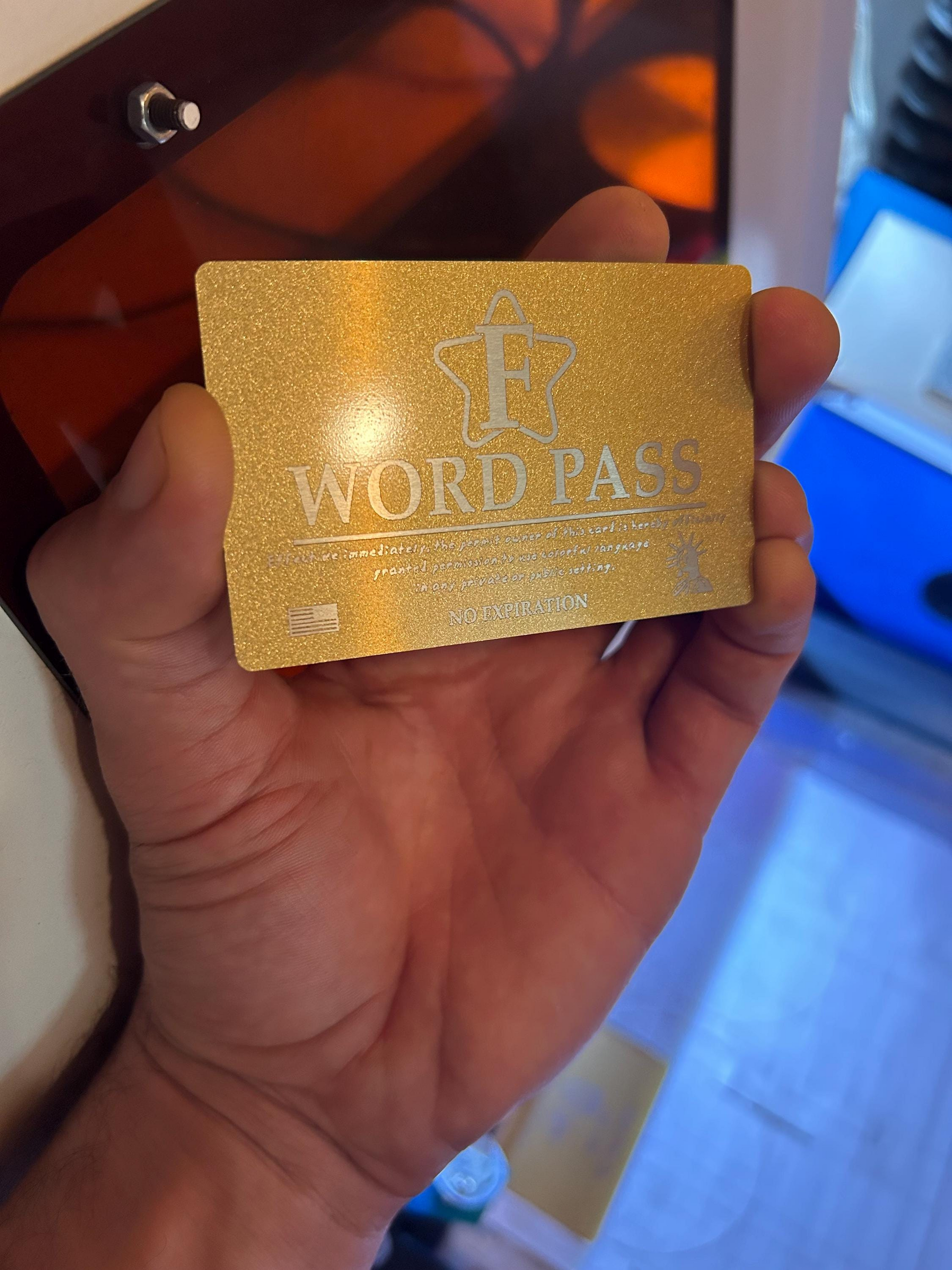 Gold/silver metal novelty gift card with personalized 'F WORD PASS' text held in a hand against a laser engraving machine background