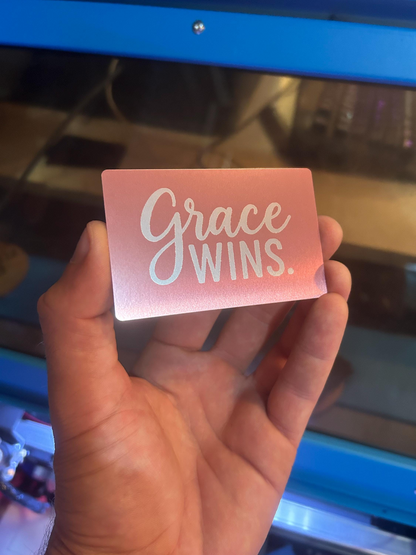 Hand holding an anodized aluminum rose gold keepsake card with 'Grace Wins' text in a laser machine setting