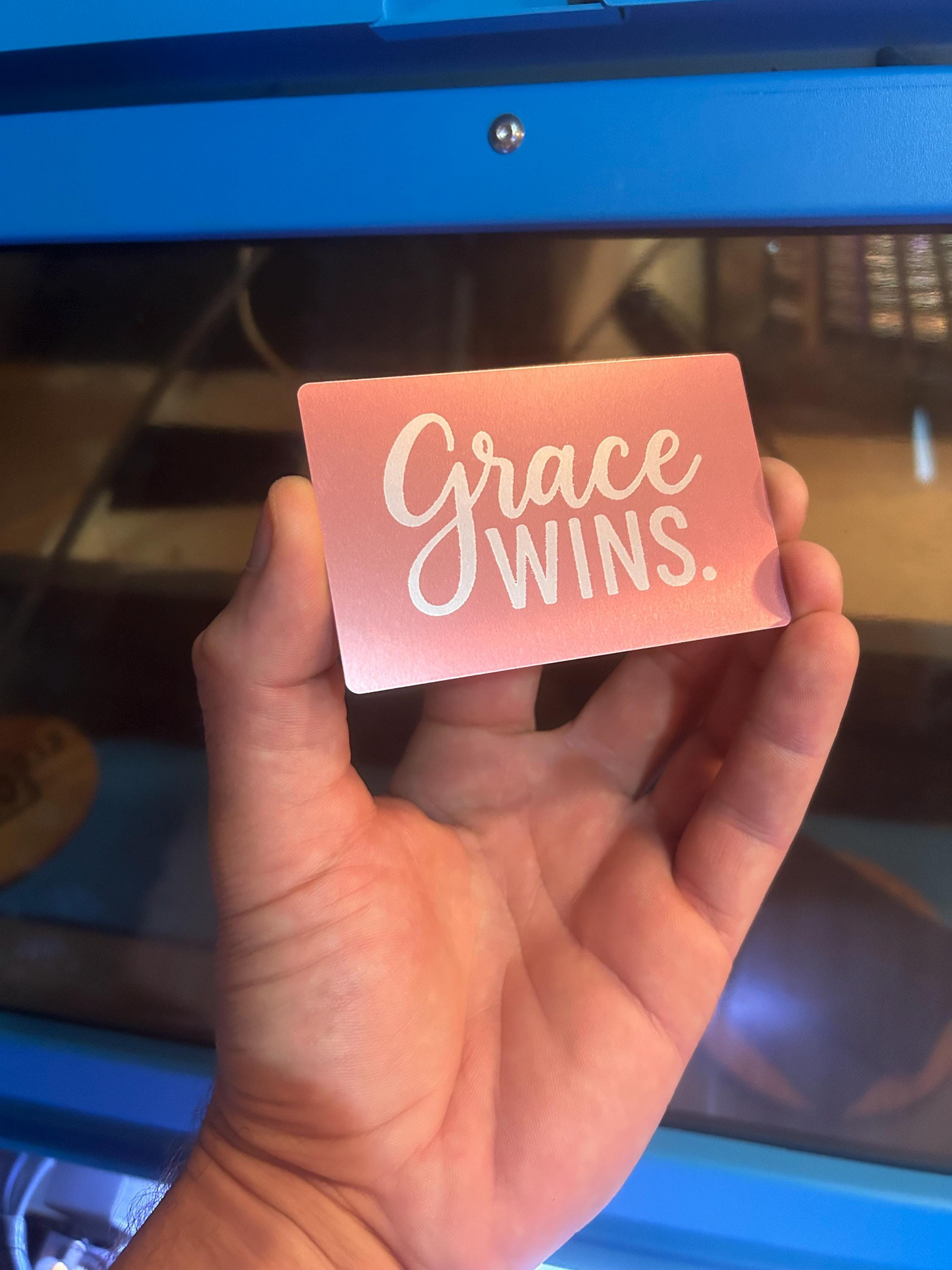 Hand holding a pink metal business card with 'Grace Wins' typographic design against a blue background