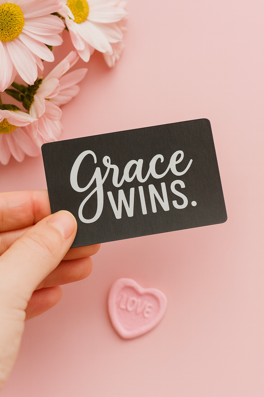 Hand holding a metal keepsake card with 'Grace Wins' text, flowers, and a pink heart on a pink background