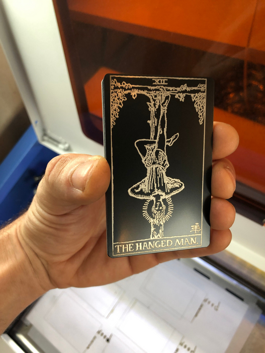 Hand holding a metal laser engraved anodized aluminum tarot card titled 'The Hanged Man' in an indoor setting.