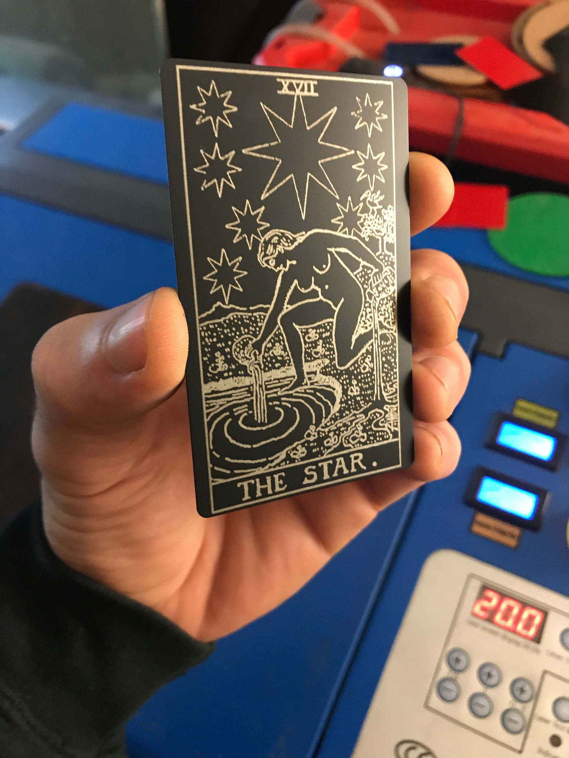 Metal business card with The Star tarot card design laser engraved on matte black anodized aluminum in front of a cnc machine