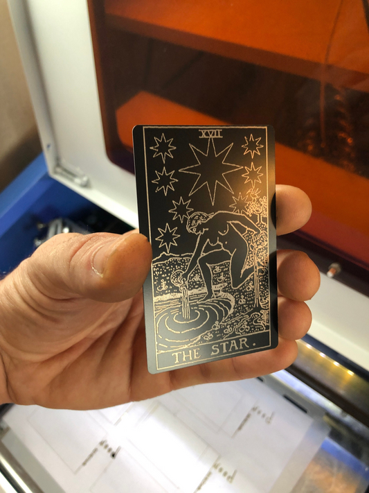 Hand holding a metal laser engraved tarot card with 'The Star' design against a blurred background