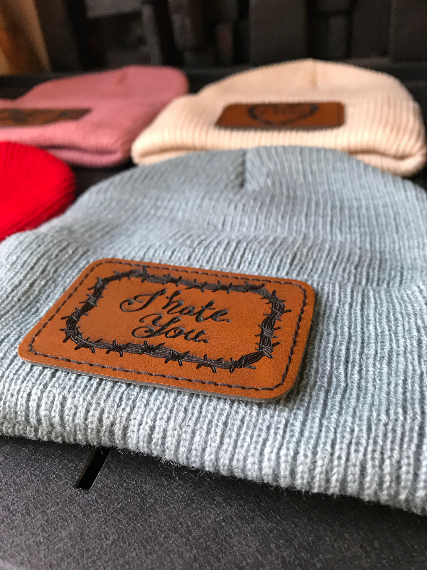 Gray beanie with a brown leather patch reading 'I Hate You' on a dark background.