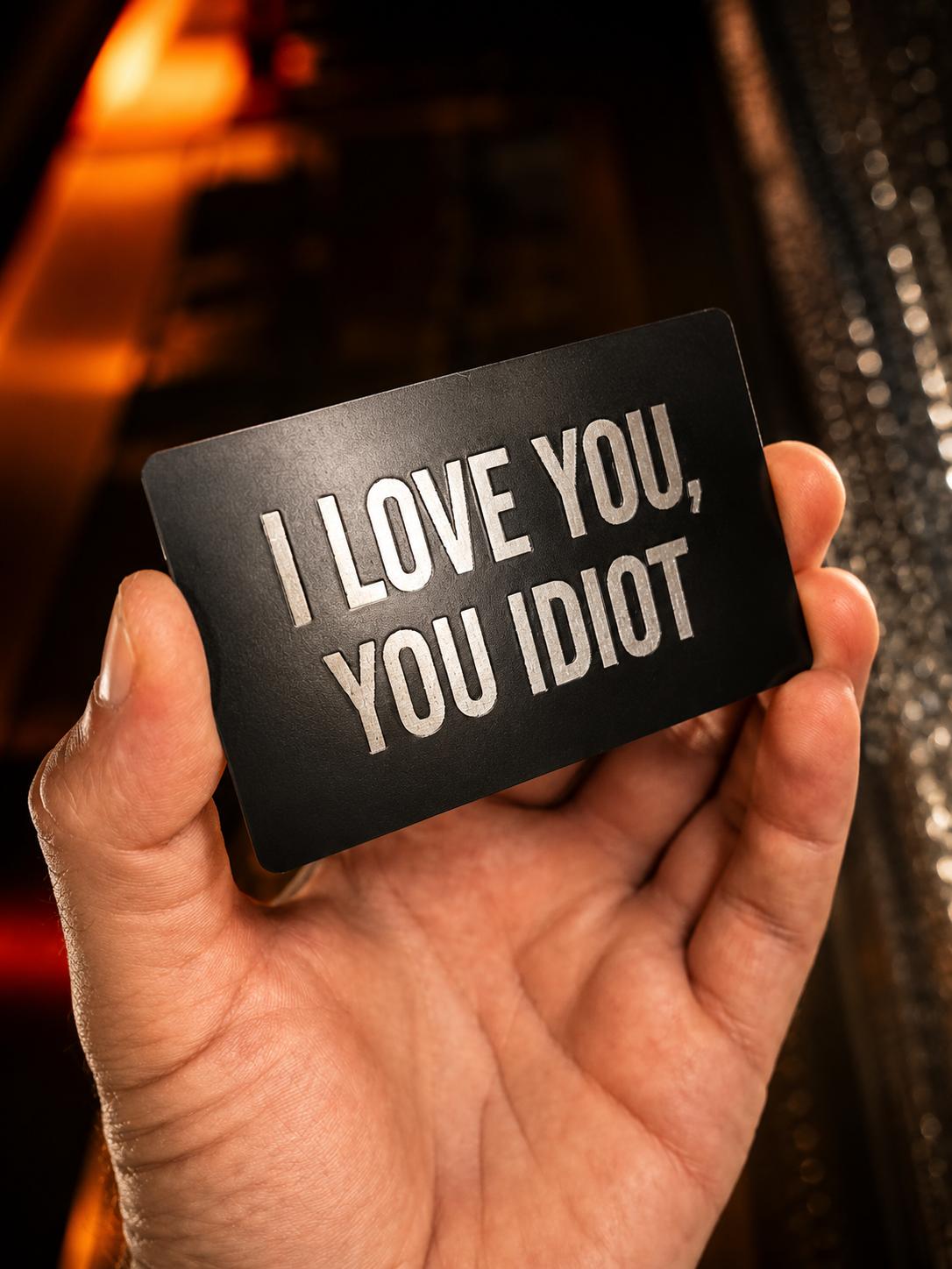 Metal keepsake funny gift card that reads "I Love you, You idiot!" laser engraved in black/silver anodized aluminum
