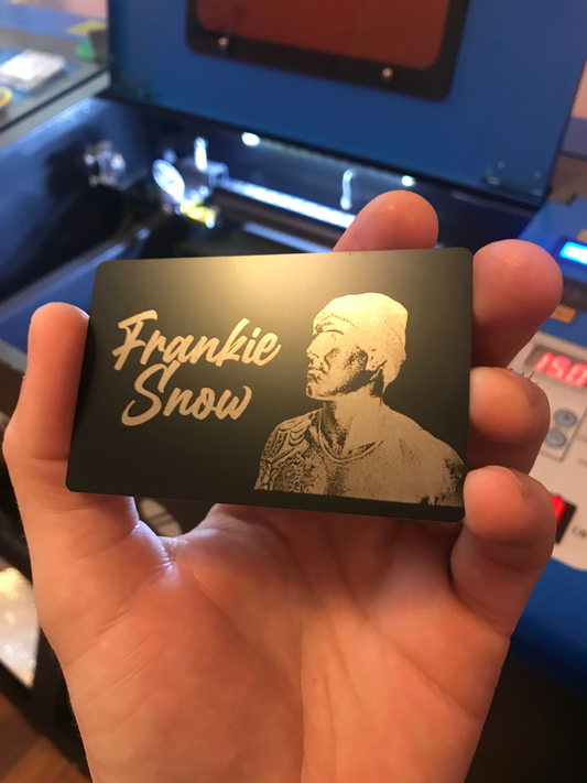 Hand holding a black/silver metal business card with engraving of a person's face and custom name text in front of a laser cutting machine.