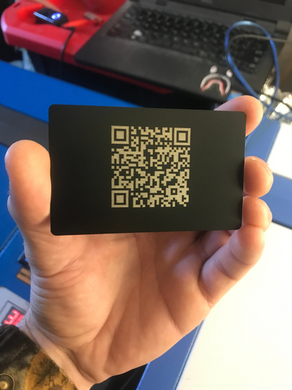 Hand holding a black/silver metal scannable business card with a QR code in front of a laptop.