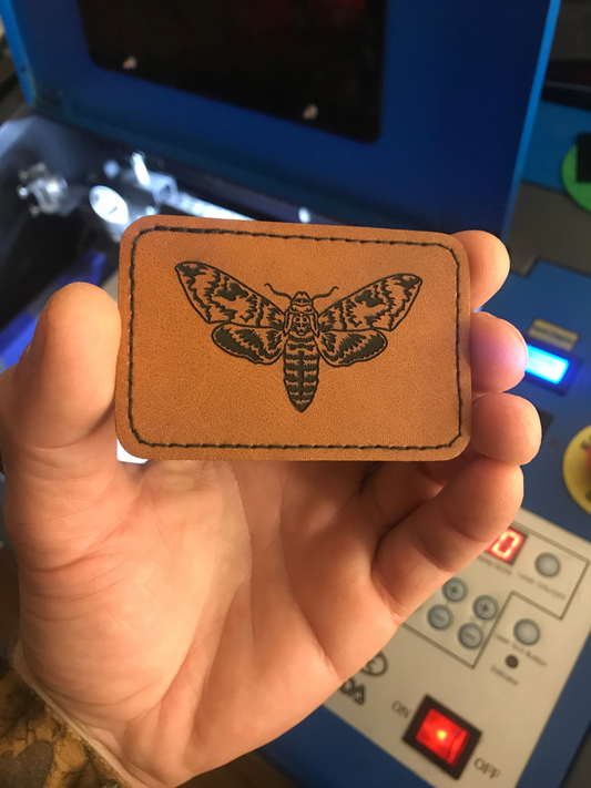 Hand holding a brown leather patch with a cicada design against a blurred background.