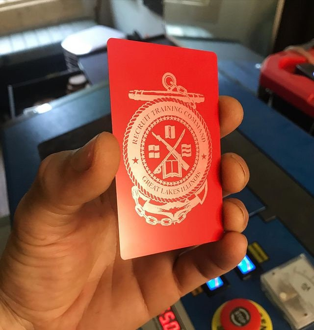 Red/silver metal business card card with a custom logo image laser engraved, held by a hand in front of a cnc machine 